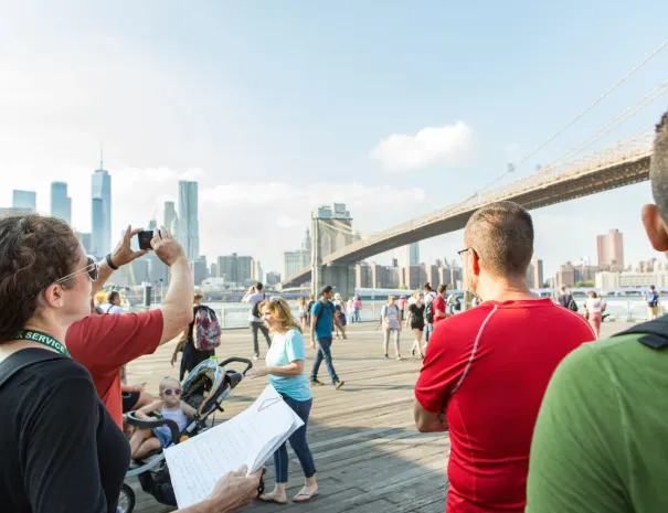 Tourists at Brooklyn Bridge Park facing the bridge A group of people standing on the Brooklyn waterfront, one taking a photo, with the Brooklyn Bridge and Manhattan skyline in the background.