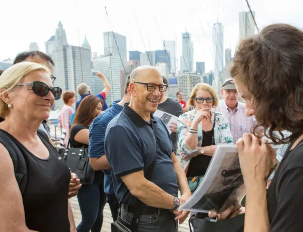 Group gathered around guide with photo on the bridge A tour guide shows a historic photo to a smiling group of visitors on the Brooklyn Bridge, with the Manhattan skyline in the background.