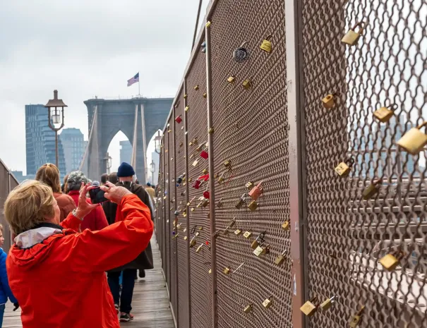Visitor photographing love locks on bridge fence A woman in a red jacket takes a photo of a fence covered in love locks on the Brooklyn Bridge, with the bridge towers and American flag in the distance.