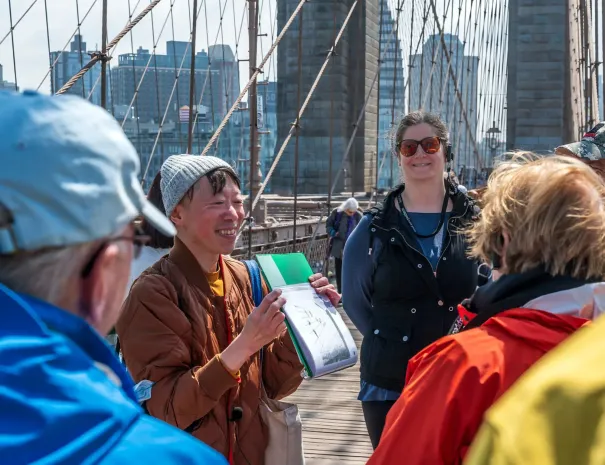 Tour guide smiling on the Brooklyn Bridge A tour guide holding papers and speaking to a small group on the Brooklyn Bridge, with suspension cables, stone towers, and city buildings in the background.