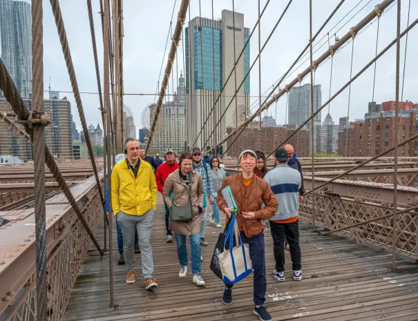 Group walking across the Brooklyn Bridge Tour participants walk along the wooden pedestrian walkway of the Brooklyn Bridge, smiling, with suspension cables and city buildings around them.