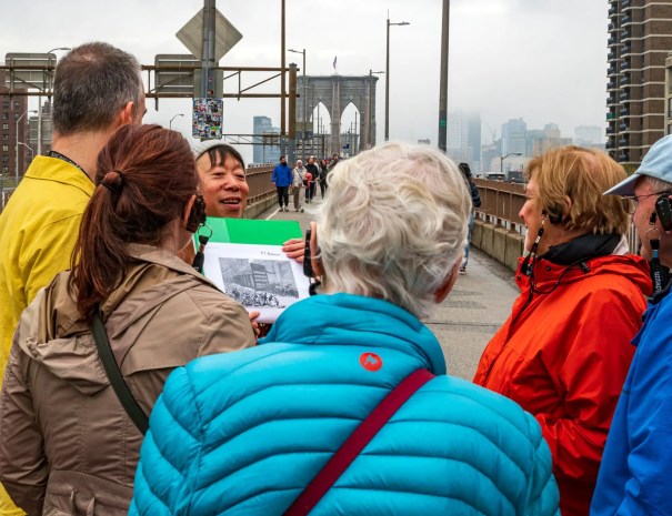 Guide showing historical image before entering bridge A guide holds up a historic illustration to a group of visitors at the entrance to the Brooklyn Bridge, with foggy skyscrapers in the background.