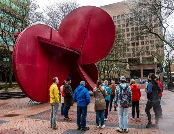 Tour group at red abstract sculpture A group of tourists gathers around a large red steel abstract sculpture in a public plaza, with brick buildings and trees in the background.