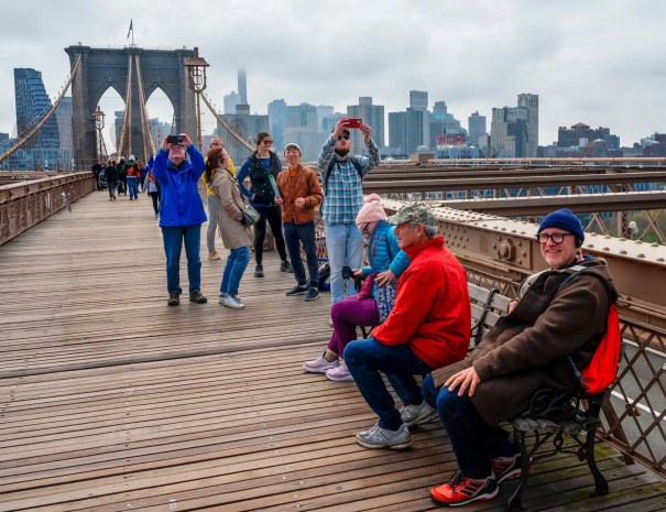 Tourists relaxing and taking photos on the Brooklyn Bridge Visitors stand and sit on benches along the wooden walkway of the Brooklyn Bridge, taking photos with the bridge towers and skyline behind them.