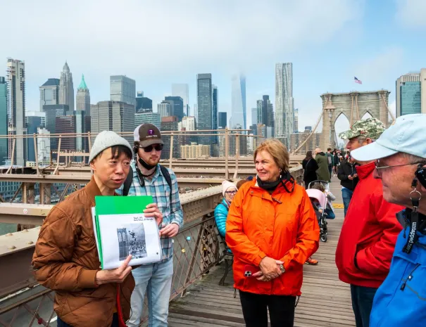 Guide explaining bridge history with skyline behind A tour guide holds an archival image while speaking to visitors on the Brooklyn Bridge, with the Manhattan skyline in the background.