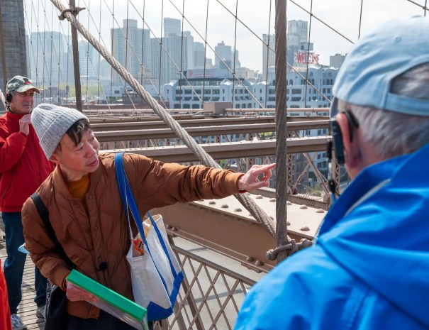 Guide pointing to bridge structure The tour guide points toward the bridge’s steel cables and beams while explaining a feature to a visitor, with buildings visible across the river.