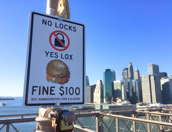 “No Locks, Yes Lox” sign on the bridge A humorous sign reading “No Locks, Yes Lox — Fine $100” with an image of a bagel, mounted on the bridge, with the Manhattan skyline and river in the background.