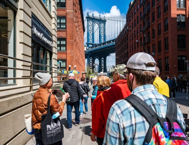 Tour group in DUMBO with Manhattan Bridge view A guide leads a group through DUMBO, Brooklyn, with the Manhattan Bridge framed between red brick warehouse buildings in the distance.