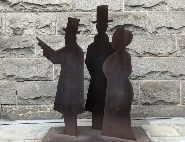 Silhouette sculpture of historical figures A metal silhouette sculpture of three Victorian-era figures in top hats and long coats, positioned against a gray stone wall.