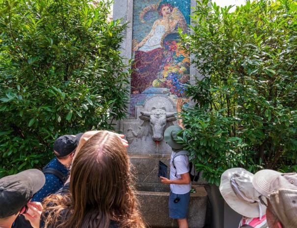 Guests gather at a granite fountain with a carved bull's head spout below a colorful glass mosaic panel, framed by dense green shrubs.