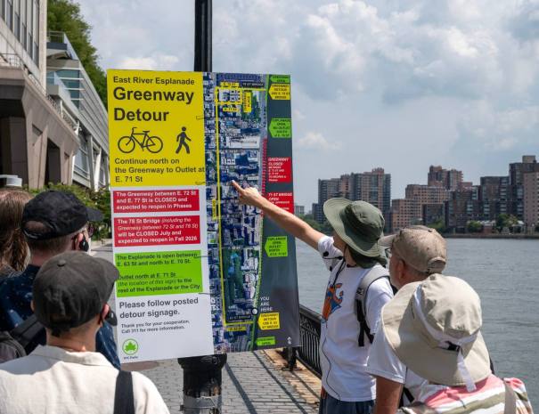 Tour group gathered around an NYC Parks "East River Esplanade Greenway Detour" sign as the guide points to the closure map.