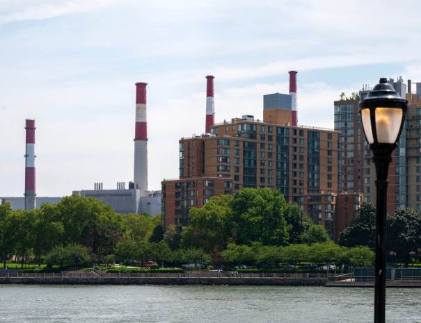 The red-and-white striped smokestacks of the Ravenswood Generating Station rise behind riverfront apartment towers and trees across the East River.