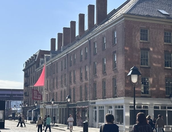 Historic red-brick warehouse building at the South Street Seaport with a red triangular flag, pedestrians walking along a cobblestone waterfront plaza