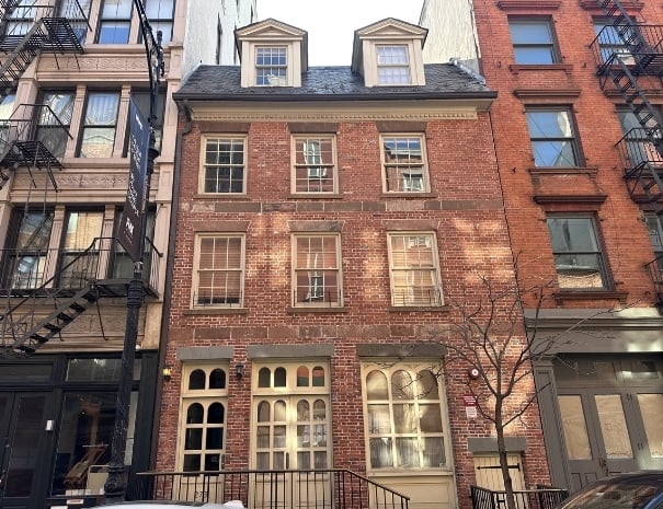 Facade of a narrow, four-story Federal-style red-brick rowhouse with a dormer window, flanked by taller neighboring buildings with fire escapes