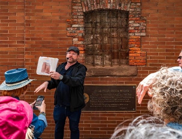 Untapped New York tour guide holding a historical illustration in front of the Sugar House Prison window plaque on a red brick wall in lower Manhattan