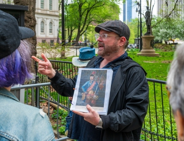 Untapped New York guide gesturing animatedly while holding a portrait of a Revolutionary War soldier during an outdoor walking tour in lower Manhattan