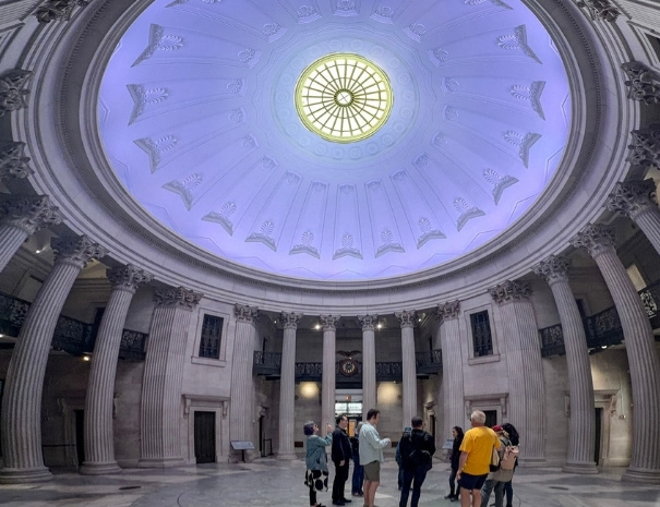 ide-angle view looking up at the illuminated coffered dome inside Federal Hall National Memorial, with an Untapped New York tour group gathered below beneath the towering Corinthian columns