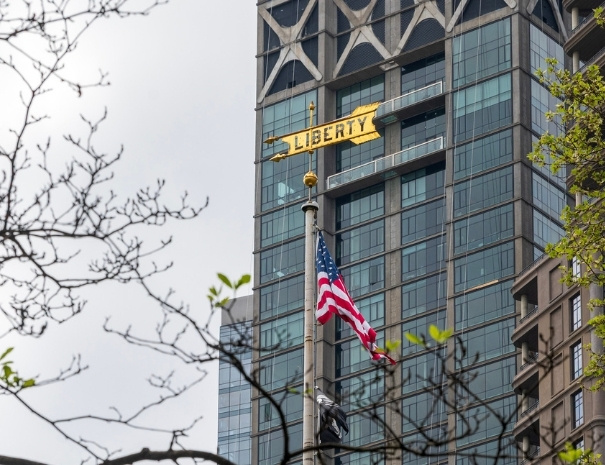 An American flag flying in front of a modern Manhattan skyscraper bearing a gold "Liberty" sign, framed by bare tree branches in lower Manhattan